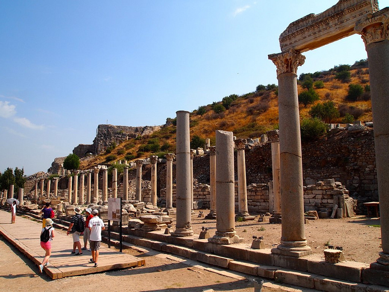 People walking alongside stone pillars at Ephesus on sunny day